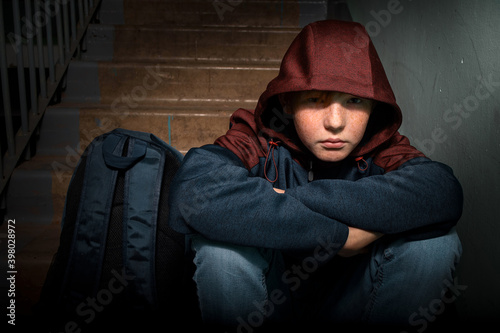 A sad red-haired teenage boy sits alone on the steps in the entrance of an apartment building.