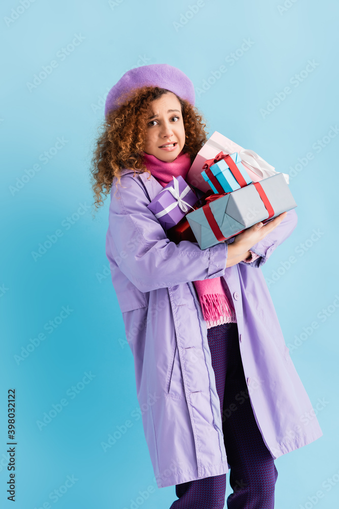 confused woman in winter coat, beret and pink knitted scarf holding bunch of christmas presents on blue confused woman in winter coat, beret and pink knitted scarf holding bunch of christmas presents on blue