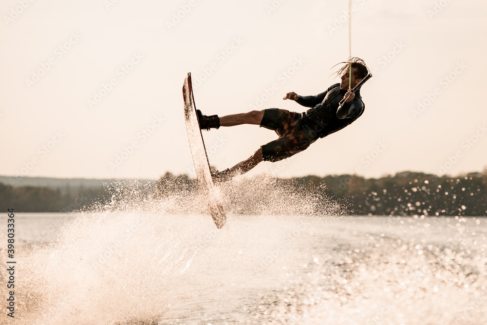 energetic muscular man holds rope and makes flips with wakeboard over splashing water