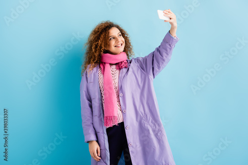 happy young woman in winter coat and pink knitted scarf holding smartphone and taking selfie on blue