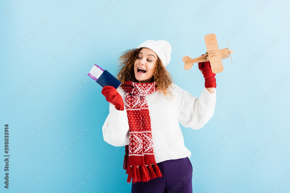 Φωτογραφία excited young woman in hat and sweater holding passport and toy plane on blue