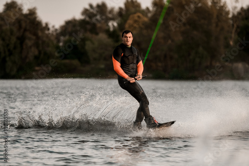 handsome man in wetsuit rides on splashed river wave on wakeboard.
