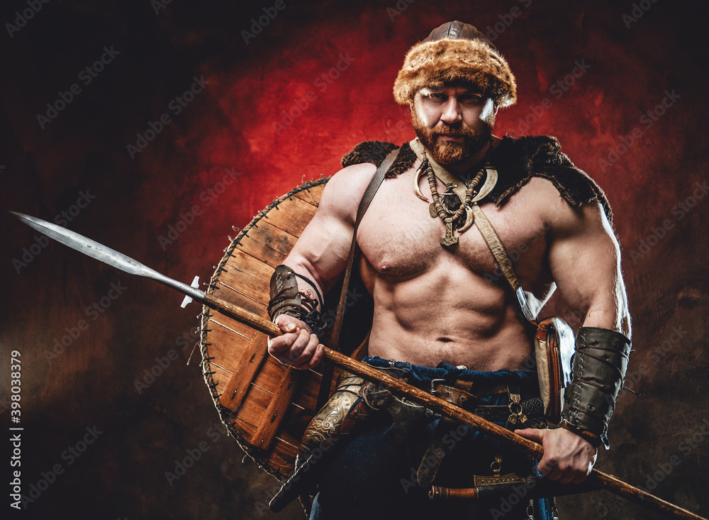 Armed with spear muscular and shirtless scandinavian barbarian with shield on his back poses in dark red background looking at camera.