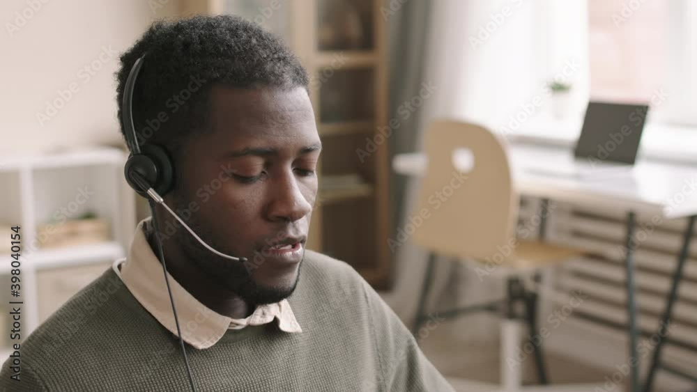 Medium shot of young African-American male call center worker wearing headset sitting at desktop and answering phone call