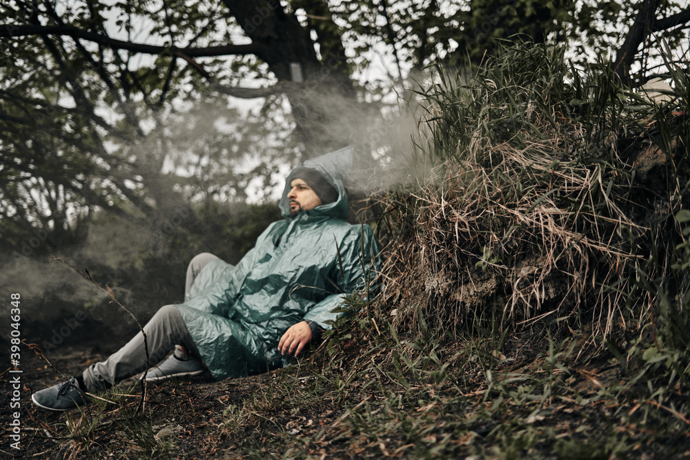 A man with a beard in a green raincoat sits on the ground in nature, the smoke from the fire is visible. Rest in cloudy weather.