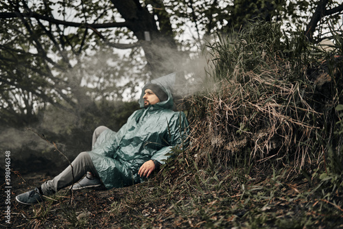 A man with a beard in a green raincoat sits on the ground in nature, the smoke from the fire is visible. Rest in cloudy weather. © Natalya Lys