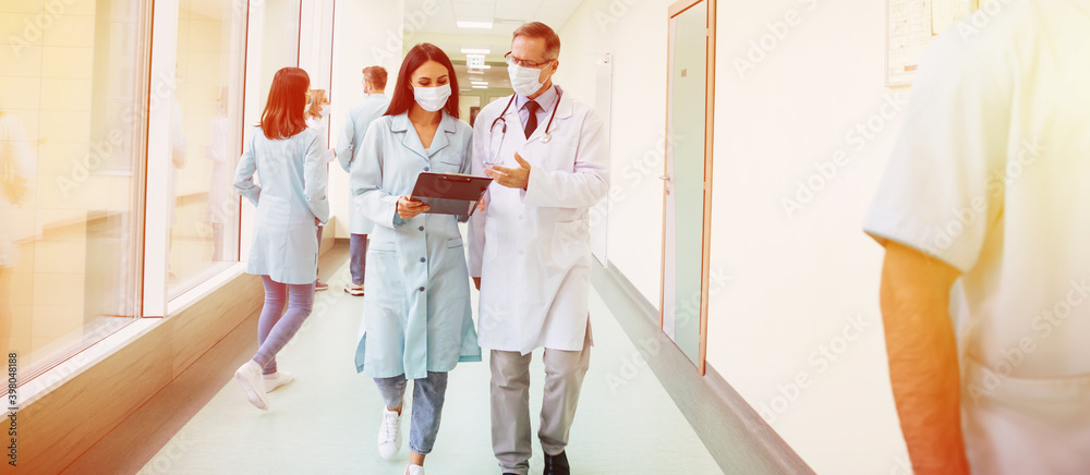 © My Ocean studio - Two confident doctors in safety medical masks have a discussion about some treatment methods during walking the hospital corridor with other colleagues around them © My Ocean studio - Two confident doctors in safety medical masks have a discussion about some treatment methods during walking the hospital corridor with other colleagues around them