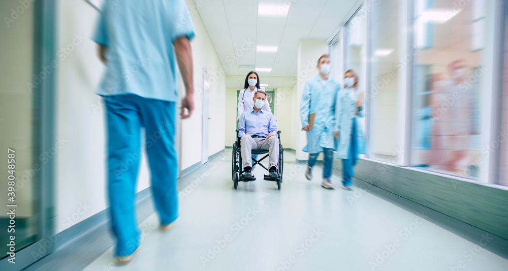 Obraz premium Ill senior man with protective safety mask on face in a wheelchair and a confident doctor in the medical mask while transporting on the hospital.