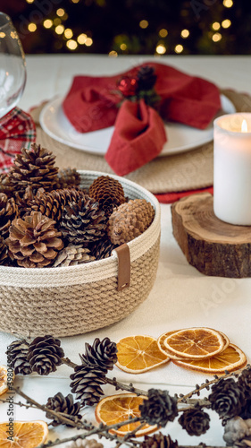 Festive holiday table vintage style with pine cones and dried fruit as decoration, a red napkin and a white table cloth in front of a decorated Christmas tree with lights