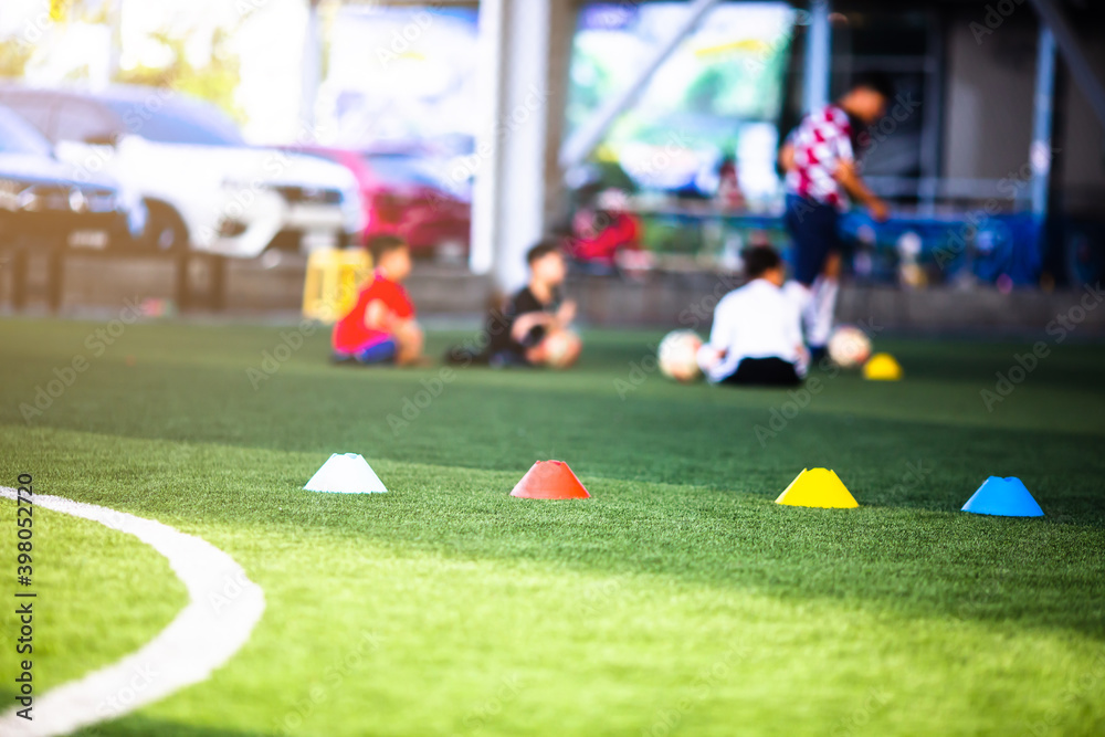 Obraz premium Selective focus to cone marker is soccer training equipment on green artificial turf with blurry kid players training background. Material for training class of football academy.