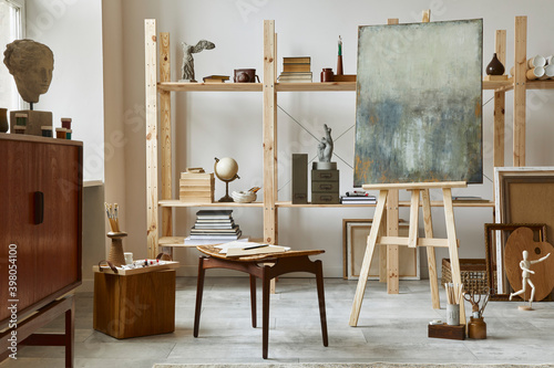 Unique artist workspace interior with stylish teak commode, wooden easel, bookcase, artworks, painting accessories, decoration and elegant personal stuff. Modern work room for artist. Template. photo