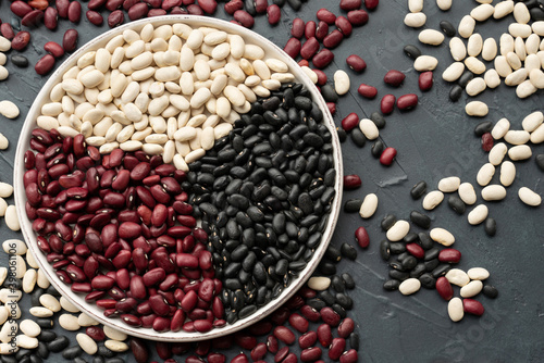 beans in white ceramic plate. bowl of beans. black bean, red kidney bean and white bean. navy bean, cannellini bean, white kidney bean on dark gray background and scattered various beans