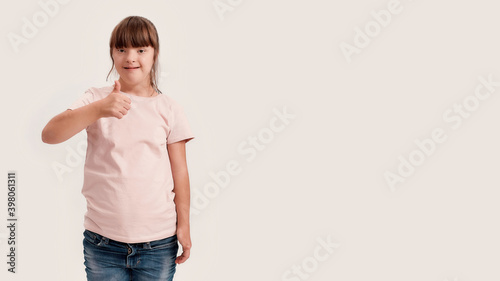 Portrait of disabled girl with Down syndrome smiling at camera, showing thumbs up with one hand while standing isolated over white background