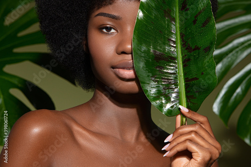 Beauty portrait of young beautiful african american woman with posing with banana leaf curly hair against green exotixc plants  background. Natural skin care concept