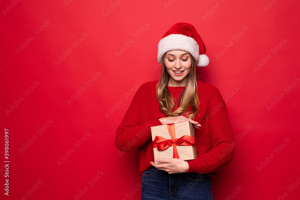Beautiful lwoman in santa hat holding giftbox in hands isolated on red background Beautiful lwoman in santa hat holding giftbox in hands isolated on red background