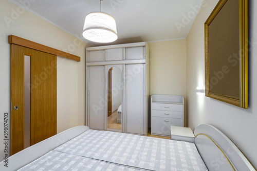 Contemporary interior of bedroom in modern flat. White furniture. Sliding wooden door. Wardrobe with mirror.