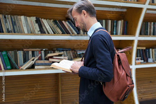 Young man with leather backpack reding book on city bookcrossing station. College student at street library book exchange point, free public bookcase.