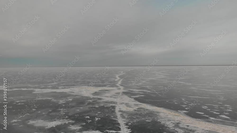 Aerial high above a frozen lake that is covered in black ice with the scattered snow drifts and long cracks. The sky is full of white and gray clouds with one patch of light blue.
Aerial high above a frozen lake that is covered in black ice with the scattered snow drifts and long cracks. The sky is full of white and gray clouds with one patch of light blue.