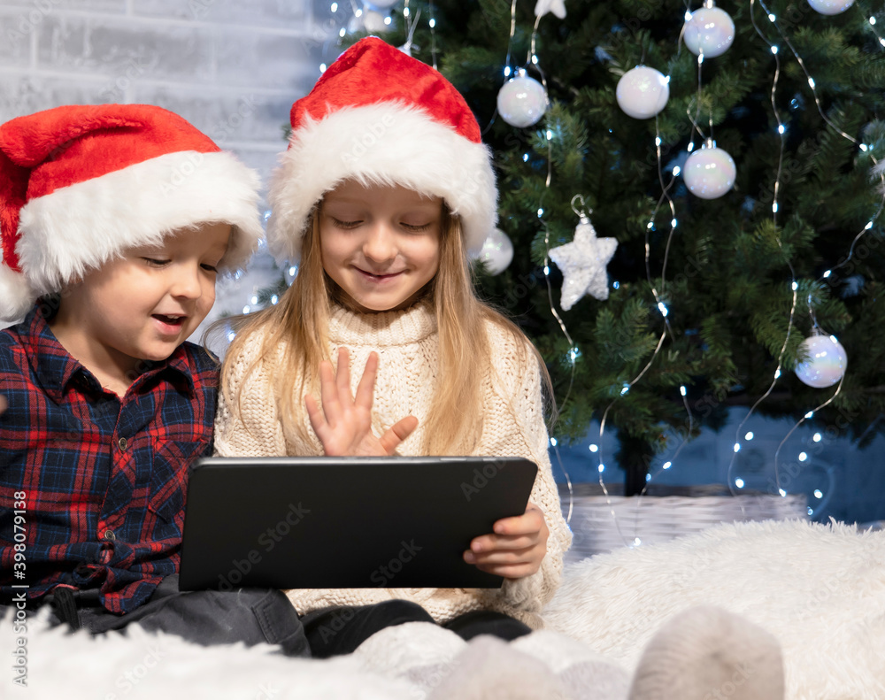 children in santaclaus hats sit near a Christmas tree on a white rug with a tablet in their hands, video call with relatives on the eve of New Year's celebrations children in santaclaus hats sit near a Christmas tree on a white rug with a tablet in their hands, video call with relatives on the eve of New Year's celebrations