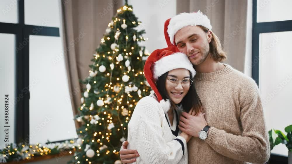 Multiracial pair in Santa hats are smiling and hugging each other standing at home against green Christmas tree Multiracial pair in Santa hats are smiling and hugging each other standing at home against green Christmas tree