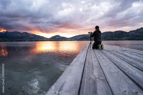 Experience nature with the hobby of outdoor photography - A man kneeling on a wooden pier of a lake adjusts his camera aiming at a gorgeous cloudy sunset in the mountains with orange water reflections