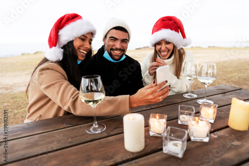 Three happy young mixed-race friends sitting outdoor in Santa Claus hat smiling making selfie with lowered Coronavirus safe protection mask with to celebrating Christmas holidays - Focus on man