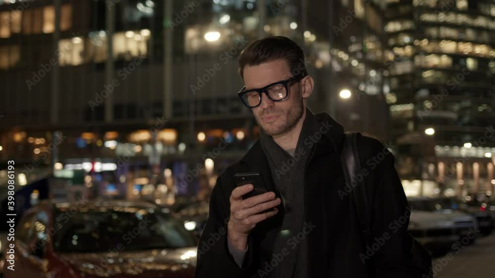Man in eyeglasses with phone in hand on background of building with lights on. Handheld night shot of caucasian man texting in phone near business building with cars Man in eyeglasses with phone in hand on background of building with lights on. Handheld night shot of caucasian man texting in phone near business building with cars