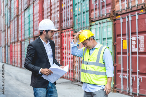 Boss business man is complaining worker by hand holds drawing or A3 paper and worker hold his hand on top of yellow hardhat like apologizing to the boss with cargo container yard background.