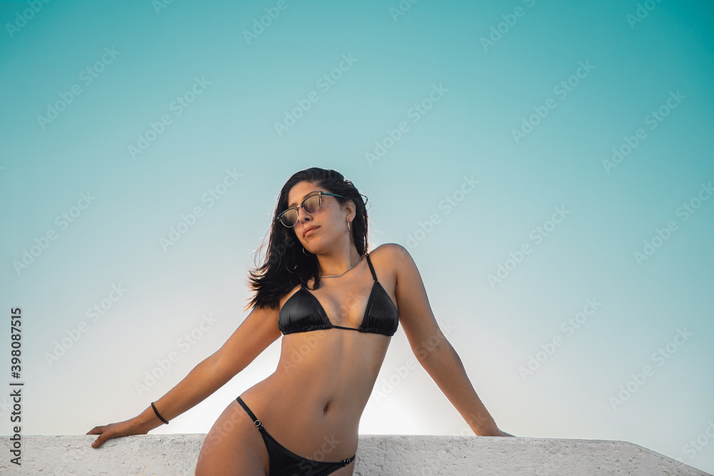 Joven modelo femenina con lentes de sol morocha bronceada en bikini negro  posando sobre una baranda blanca en un atardecer de cielo cyan Stock Photo  | Adobe Stock