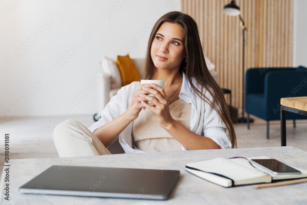 Beautiful brunette girl sits at table in front of laptop, holds cup of coffee with her hands. Woman is drinking coffee Beautiful brunette girl sits at table in front of laptop, holds cup of coffee with her hands. Woman is drinking coffee
