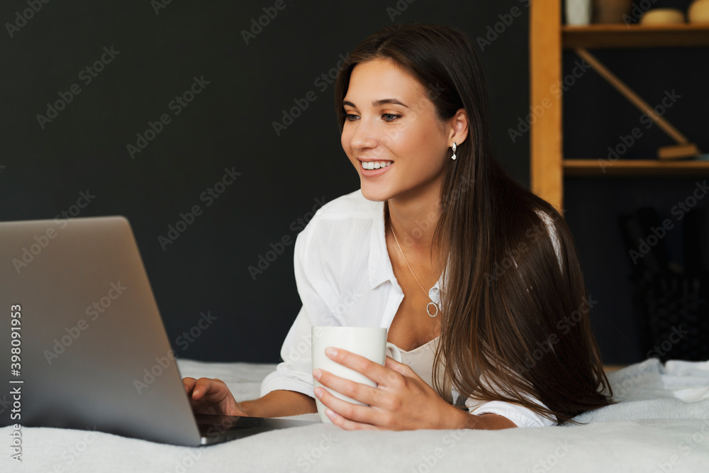 Millennial girl is sitting on bed in white shirt holding cup of coffee in her hand. Beautiful brunette woman uses laptop Millennial girl is sitting on bed in white shirt holding cup of coffee in her hand. Beautiful brunette woman uses laptop