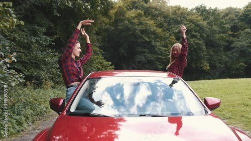 Girfriends enjoying time together. Two female friends sitting from ides of the red car haing hand up in the air while riding in the car. Weekend activities. photo