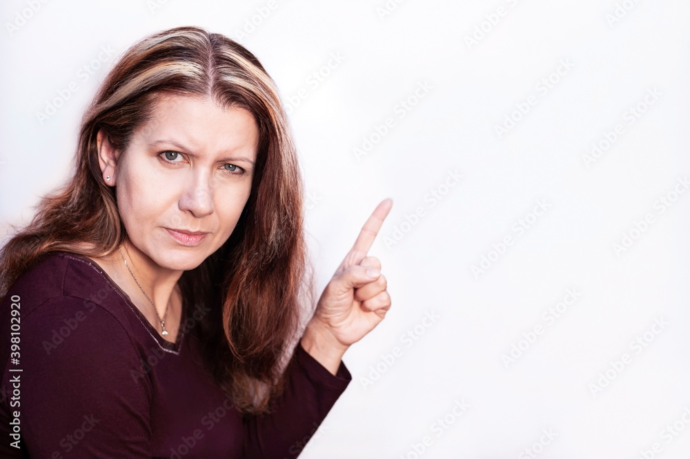 Woman on a light background looks ahead at the camera and points a finger at an empty space next to her, a place for text. The woman has a strict, slightly angry face Woman on a light background looks ahead at the camera and points a finger at an empty space next to her, a place for text. The woman has a strict, slightly angry face