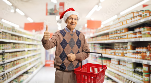 Elderly man with a shopping basket in a supermarket showing a thumb up sign amd wearing a santa claus hat photo