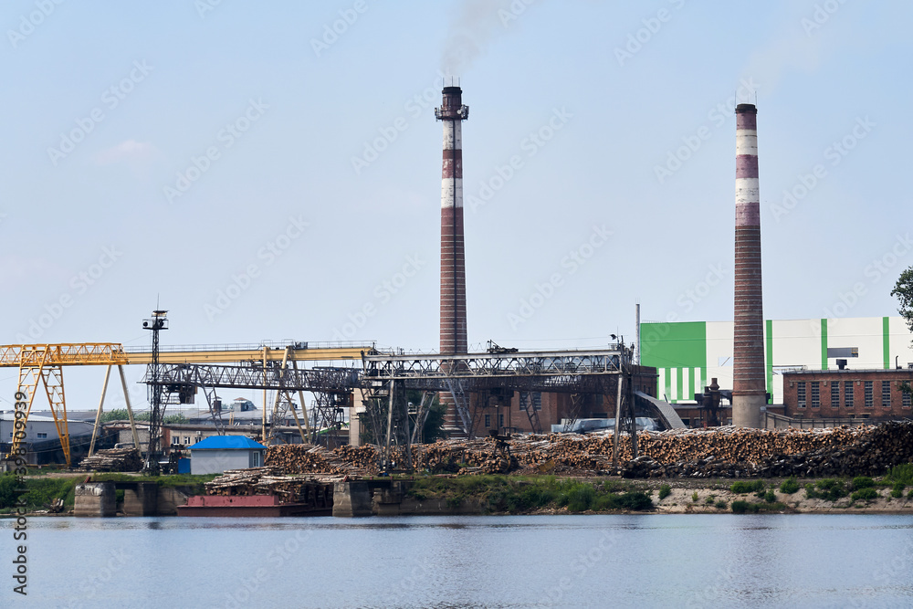 industrial landscape, pulp and paper mill with stacks of logs on the river bank
