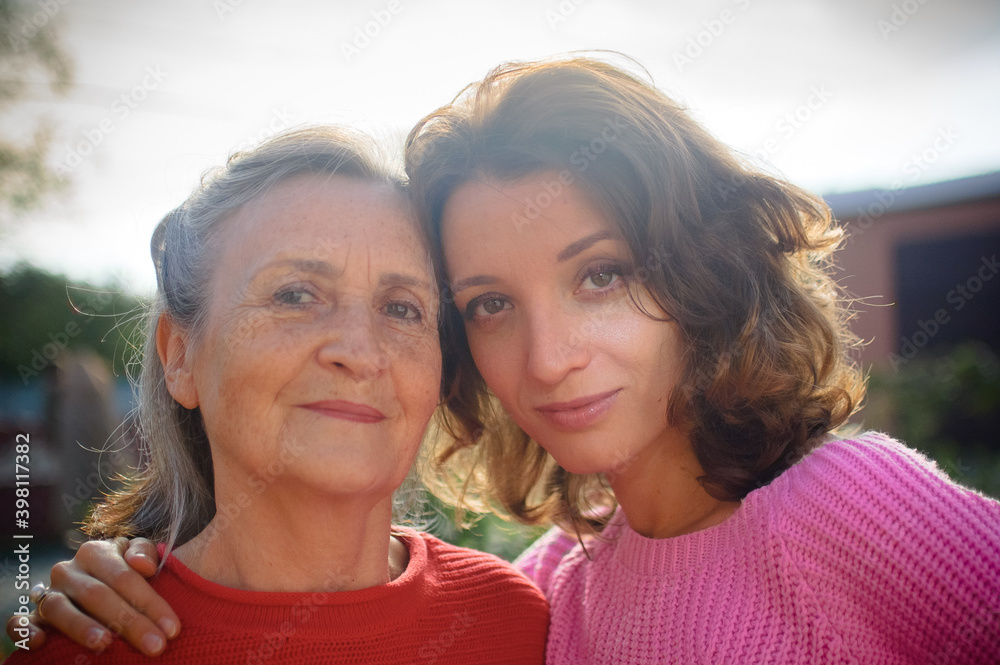 Obraz premium Senior mother with gray hair with her adult daughter looking at the camera in the garden and hugging each other during sunny day outdoors