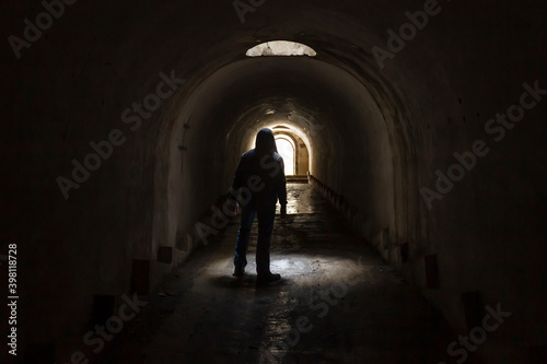 A hooded man walking towards a lighted exit from a dark, gloomy underground corridor. Light at the end of the tunnel leading to the exit to freedom.