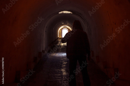 A hooded man walking towards a lighted exit from a dark, gloomy underground corridor. Light at the end of the tunnel leading to the exit to freedom. © Сергей Рамильцев