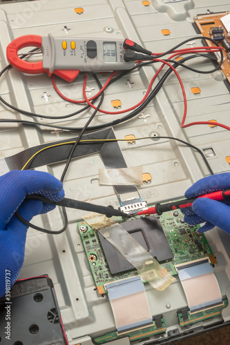 Gloved hands of an electrician engineer hold multimeter probe. Repair and diagnostics of printed circuit board of TV in service center. Selective focus