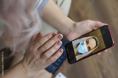 Young woman using smartphone for conversation with girlfriend via video chat at home, Long-Distance Relationship. On the phone screen a girl in a mask with a heart drawn in the color of the LGBB