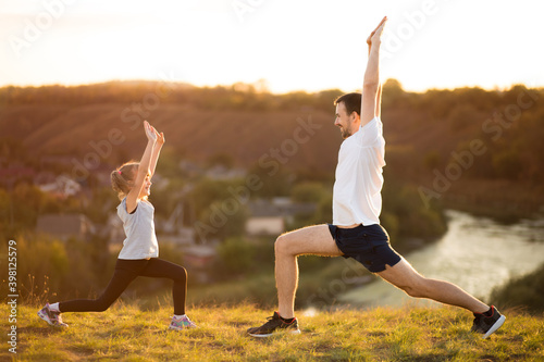 Father and cute daughter doing yoga outside in bright sunny day, lovely emotions. together at park. Family stretching after sport. Healthy habits