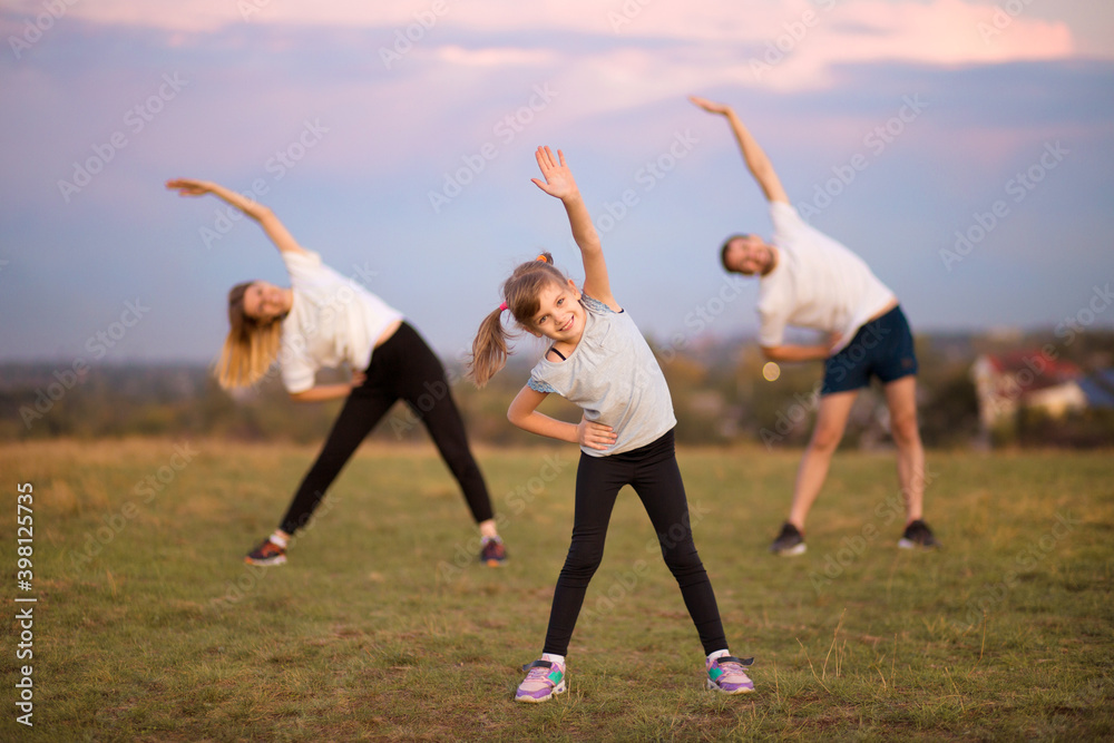 © Svetlana - Parents instill sports habits in their child by example, focus on child. Happy sporty family doing stretching exercises, practices yoga outdoor. Mom dad and daughter doing sport exercises