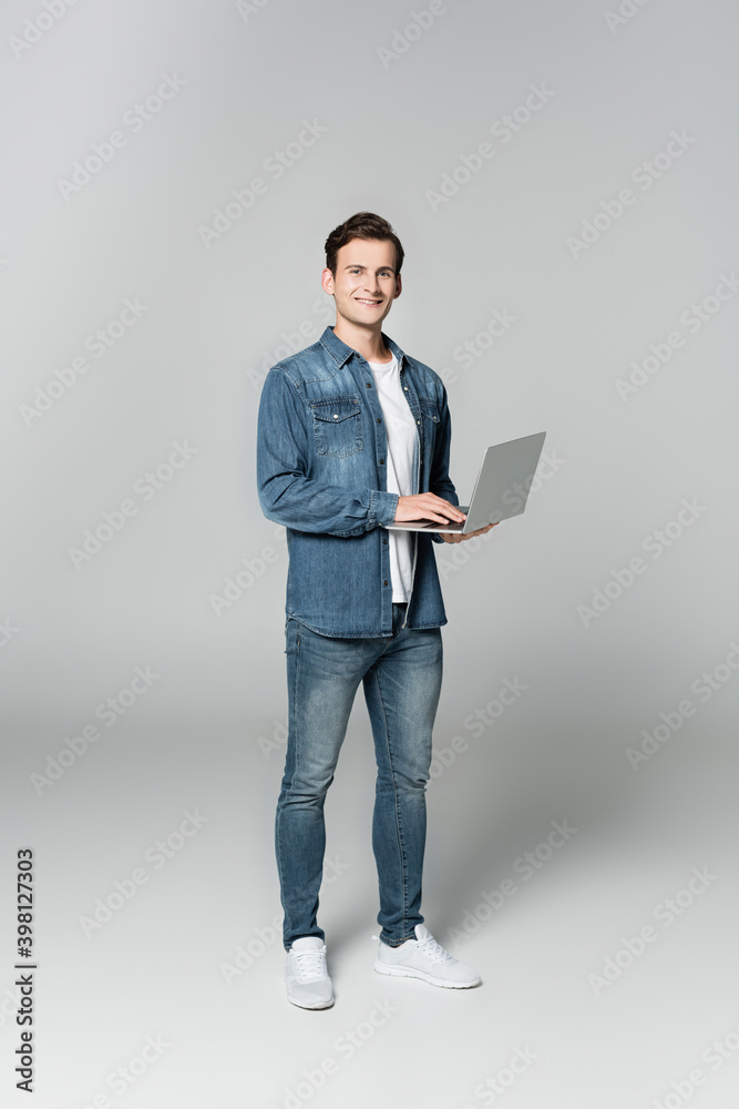 Cheerful man looking at camera while holding laptop on grey background