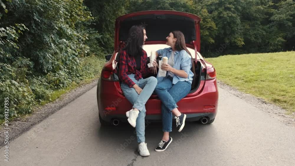 Two teenage girls sitting in a car's trunk and eating fast food before a road trip. Eating in car. Friendship. Two teenage girls sitting in a car's trunk and eating fast food before a road trip. Eating in car. Friendship.