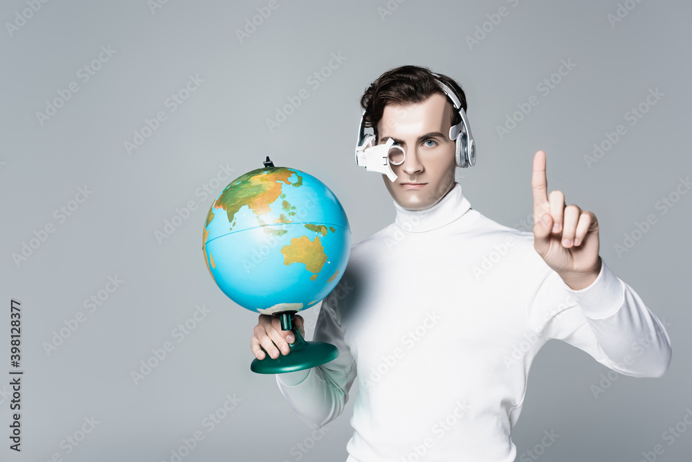 Cyborg in headphones holding globe while pointing up with finger isolated on grey Cyborg in headphones holding globe while pointing up with finger isolated on grey