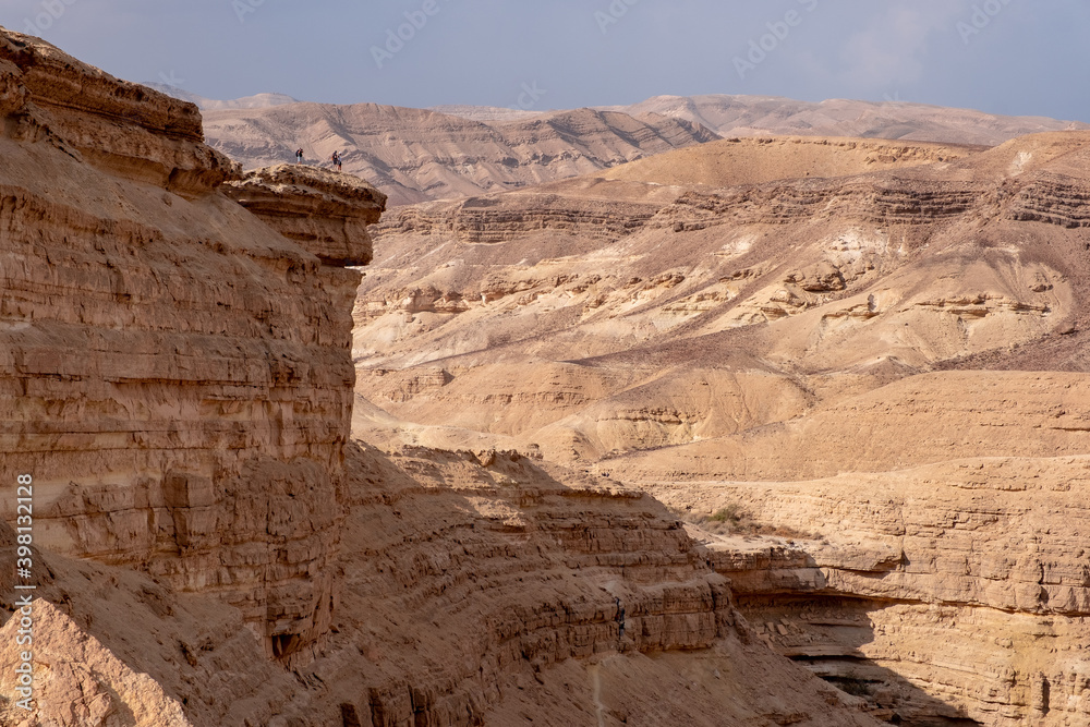 Fototapeta premium Desert landscape with sandy hills and high cliff above dry riverbed Hatira, Negev desert, Israel. Travelers standing on the cliff viewing the panorama of picturesque mountain landscape.