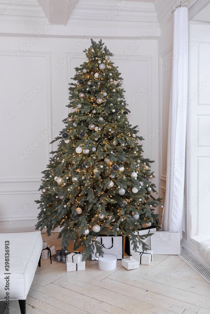 New Years interior, fir tree in green tones with lights and white, silver balls near window, light curtains. Christmas white living room interior, gifts in packaging. Location for photo shoot