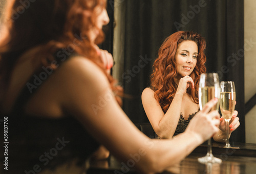 beautiful red-haired girl holding a glass of champagne in her hands and looking in the mirror