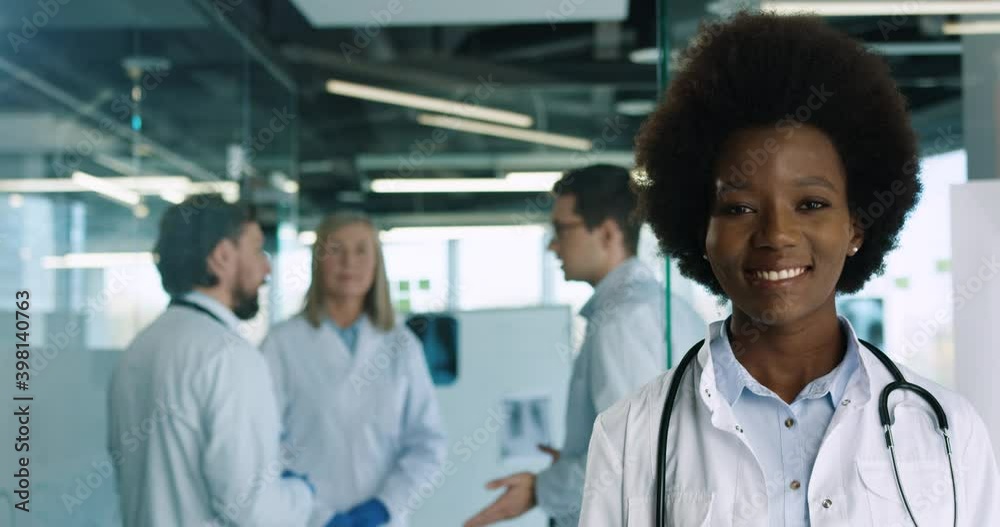 Close up portrait of young African American female professional doctor standing in hospital and smiling at camera. Caucasian doctors speaking on background. Healthcare concept Close up portrait of young African American female professional doctor standing in hospital and smiling at camera. Caucasian doctors speaking on background. Healthcare concept