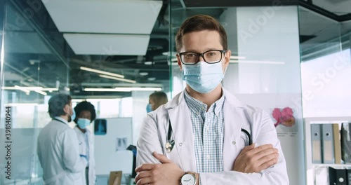 Close up of Caucasian joyuful handsome young male professional medical practitioner in glasses wearing mask standing in clinic and looking at camera. Mixed-races doctors on background. Covid19 concept photo